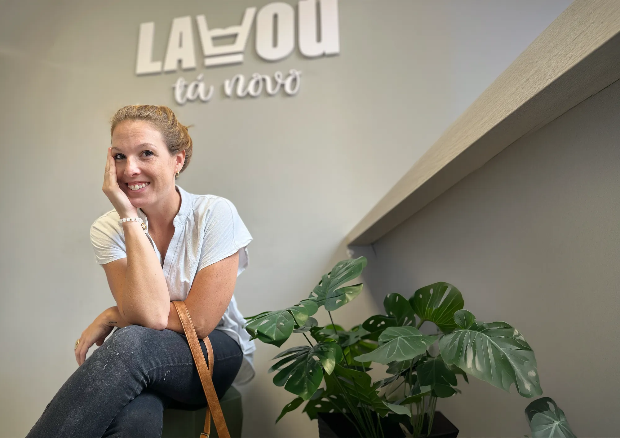 Moradora sorrindo sentada em uma lavanderia Lavou tá Novo, com o logo na parede